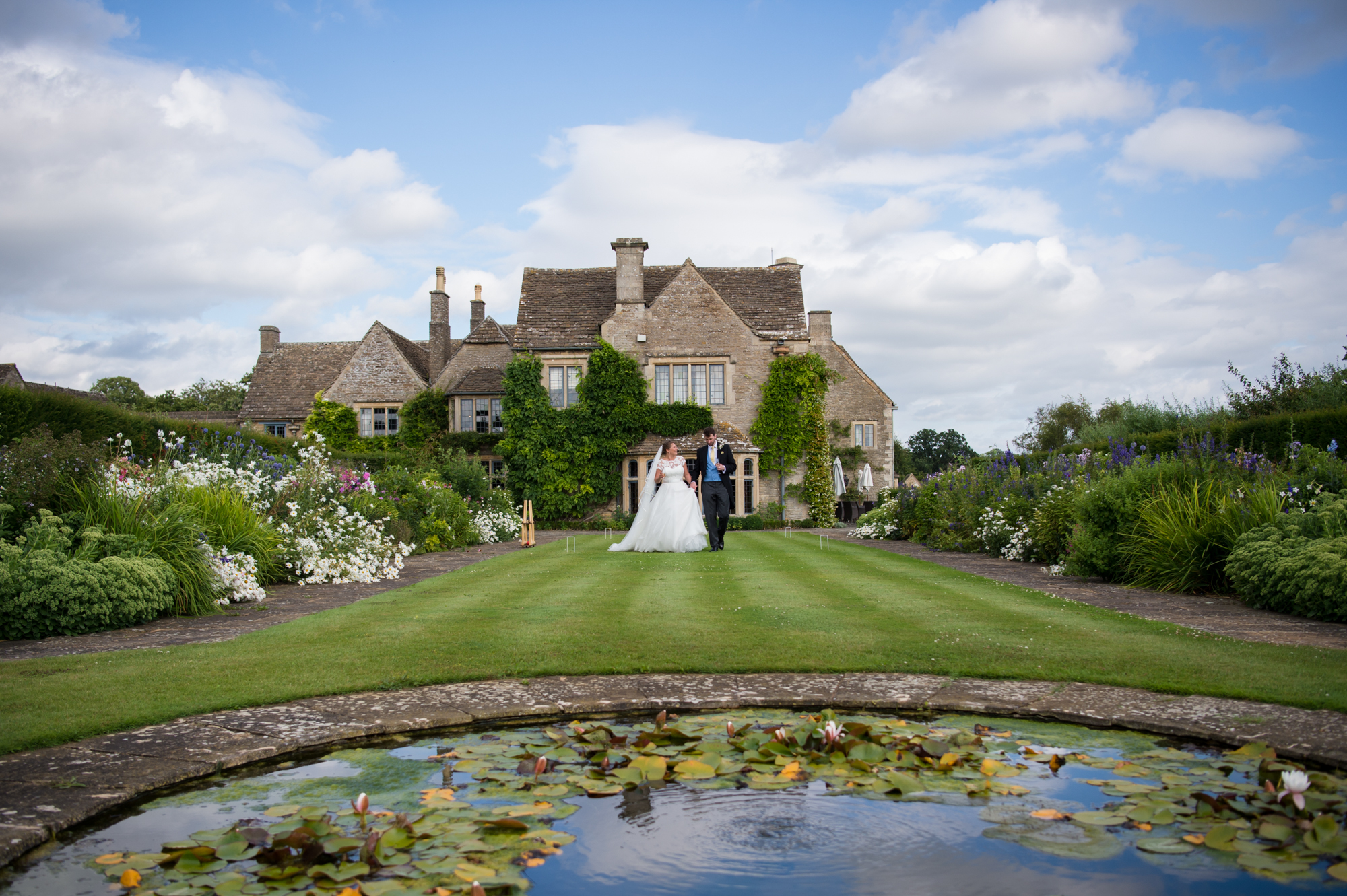Bride and groom walking in the gardens of Whatley Manor with the wedding venue in the background